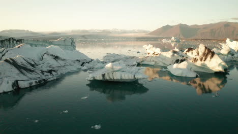 Sunlight-On-Icebergs-During-Golden-Hour-Sunrise-In-Jokulsarlon-Glacial-Lake-In-South-Iceland---orbiting-drone-shot