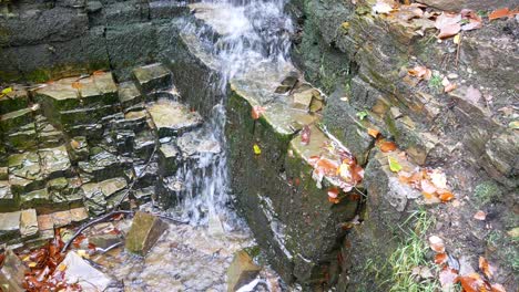 Herbstlicher-Wasserfall-Aus-Fließendem-Stein,-Der-In-Ein-Klares-Wasserbecken-Aus-Rotgoldenen-Herbstblättern-Kaskadiert