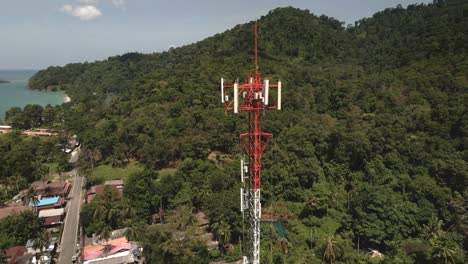 Aerial-slow-orbit-drone-shot-of-a-red-and-white-telecommunications-tower-on-a-tropical-Island-in-Thailand-with-jungle,-sea-view-and-Islands