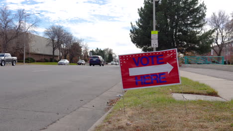 Vote-Here-Sign-Arrow-Pointing-Left-with-People-Driving-Cars-in-Background,-Wide