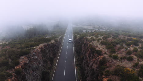 Mystic-aerial-tilt-down-showing-car-driving-on-asphalt-rural-road-into-mist-and-clouds-in-the-morning