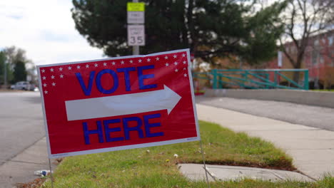 Vote-Here-Sign-Pointing-Right-with-People-Driving-Cars-and-Walking-in-Background,-Close-Up