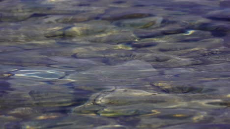 Flüssige-Textur-Des-Seewassers,-Das-Sonnenlicht-Am-Ufer-Mit-Kieselsteinen-Reflektiert