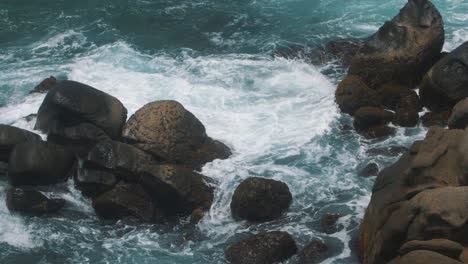 Slow-Motion-shot-of-blue-water-and-waves-crashing-against-a-stack-of-rocks-on-a-cliff,-taken-in-Tayrona-Park,-Colombia