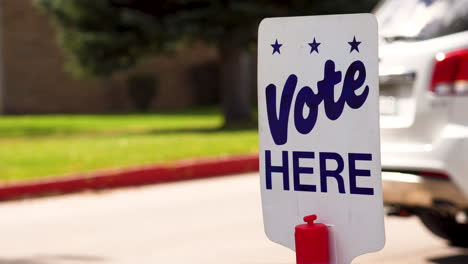 Blue-Vote-Here-Sign-Close-up-with-People-Driving-Cars-Walking-in-the-Background