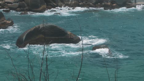 Slow-motion-zoomed-shot-of-waves-of-blue-water-crashing-against-a-rock-in-Tayrona-Park,-Colombia