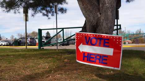 Vote-Here-Sign-Pointing-Left-with-Person-Sitting-in-Car-in-Parking-Lot-Background