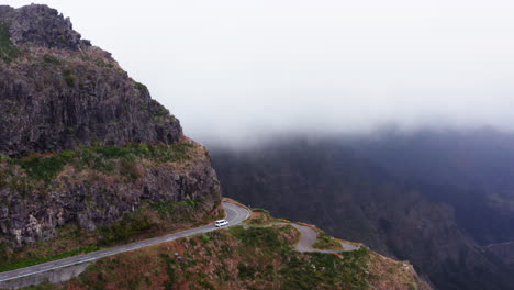 Aerial-shot-of-white-car-driving-on-dangerous-road-in-the-mountains-during-heavy-fog