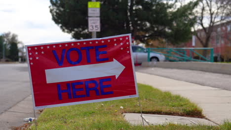 Vote-Here-Sign-Pointing-Right-with-Person-Driving-Car-Past-and-Walking-in-Background,-Close-Up