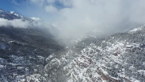 Aerial-view-of-clouds-above-white-snow-capped-hills-and-Ouray,-small-valley-town-in-southwestern-Colorado-USA-on-sunny-winter-day,-drone-shot