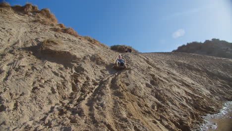 Woman-sledging-down-sand-dune-having-fun-at-Crantock-Beach,-wide-shot