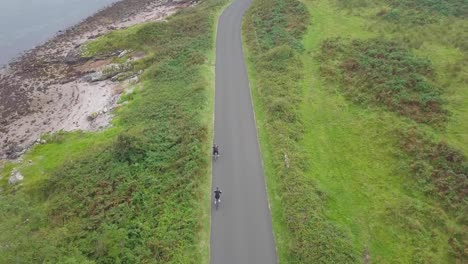 Two-People-Cycling-On-A-Bike-And-Coming-To-A-Halt-On-The-Road-Between-The-Green-Meadows,-Scotland-on-a-cloudy-day