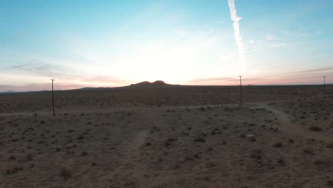 Flying-low-over-the-Mojave-Desert-landscape,-under-the-power-lines-and-over-a-dirt-road-towards-two-mountains-at-sunrise