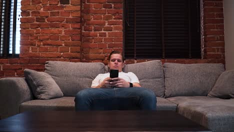 Der-Junge-Ernst-Wirkende-Mann-Sitzt-Auf-Der-Couch-Im-Wohnzimmer-Und-Schreibt-Eine-Nachricht-Mit-Seinem-Smartphone,-Frontalaufnahme