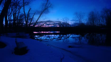Time-lapse-of-little-red,-arched,-steel-truss-bridge-over-a-river-at-dusk-with-reflections-in-the-water-and-cars-driving-by-in-small-town-America