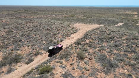 Aerial-track-shot-of-4x4-safari-vehicle-driving-on-hilly-desert-during-sunlight,-one-person-walking