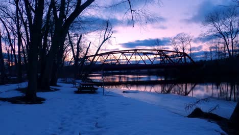 Walking-along-a-riverbank-in-a-public-green-space-park-at-sunset-during-winter-with-snow-on-the-ground,-benches-and-picnic-table,-and-a-small,-red,-trussed-arched-steel-bridge