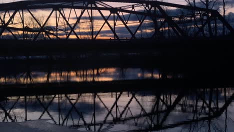 A-little-red,-arched,-steel-truss-bridge-over-a-river-at-dusk-with-reflections-in-the-water-and-cars-driving-by-in-small-town-America