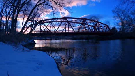 Time-lapse-of-little-red,-arched,-steel-truss-bridge-over-a-river-at-sunset-with-reflections-in-the-water-and-cars-driving-by-in-small-town-America