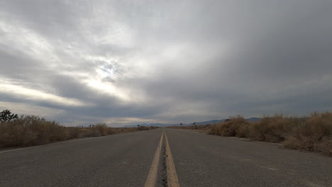Mirando-Hacia-Abajo-Una-Carretera-Vacía-En-El-Desierto-De-Mojave-Con-Un-Paisaje-Nublado-Tormentoso-Sobre-La-Cabeza---Lapso-De-Tiempo-De-Puesta-De-Sol-De-ángulo-Bajo