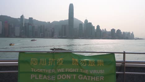 Blick-Auf-Die-Skyline-Der-Insel-Hongkong-Von-Der-Victoria-Harbour-Waterfront-Aus,-Da-Ein-Banner-Die-Öffentlichkeit-Daran-Erinnert,-Versammlungen-Zu-Vermeiden,-Um-Die-Ausbreitung-Des-Coronavirus-In-Hongkong-Zu-Verhindern