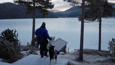 Rear-View-Of-Man-With-Dog-Walking-Down-The-Stairs-Carrying-Mat-And-Fur-Rug-Overlooking-Frozen-Lake-In-Forest-Hills-Near-Trondheim,-Norway