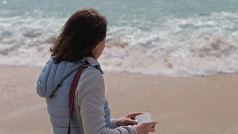 SLOW-MOTION-woman-in-warm-clothes-takes-pictures-of-waves-white-with-foam-cover-the-wet-sand-of-the-beach-on-her-phone,-Portugal