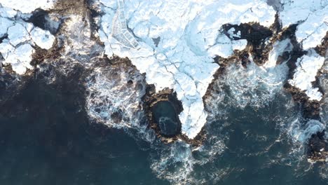 Top-down-aerial-at-Brimketill-rock-pool-with-coast-covered-in-snow,-Iceland