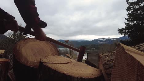 Close-up-slow-motion-of-axe-hitting-log-round-with-pieces-flying-with-lake-and-mountains-in-background
