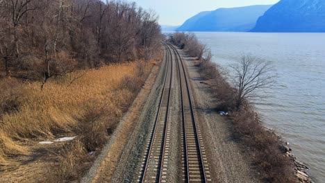 Aerial-drone-footage-down-railroad-train-tracks-in-a-river-valley-with-mountains-and-water-in-early-spring