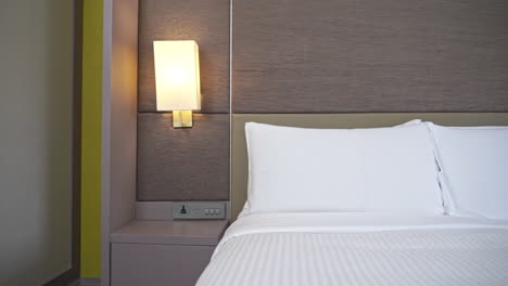 Slow-pan-right-on-classic-hotel-made-up-bad-with-high-mattress-and-turned-on-lightt-on-lamp