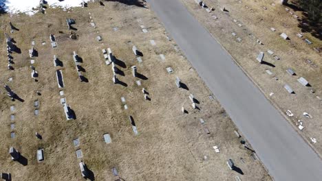 Drone-looking-down-at-a-cemetery-with-gravestones-flying-straight-a-few-seconds-then-panning-or-whipping-to-the-left-before-coming-to-a-stop,-on-a-sunny-winter-day-with-patches-of-snow-on-the-ground
