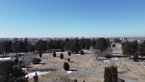 Drone-smoothly-flying-over-dead-trees-in-a-cemetery-looking-ahead-to-reveal-gravestones-on-dead-grass-with-patches-of-snow-on-a-clear-winter-day