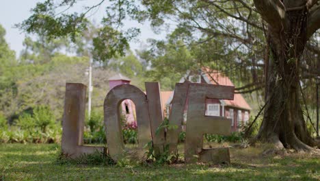 Metal-letters-forming-word-LOVE-standing-in-the-shadow-of-tree-near-German-style-house-in-background-on-a-bright-sunny-day