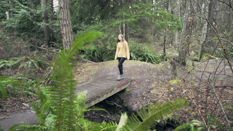A-young-woman-wearing-a-bright-yellow-jacket-walks-over-a-small-bridge-in-a-green-mossy-forest,-wide-panning-shot-from-behind-a-fern