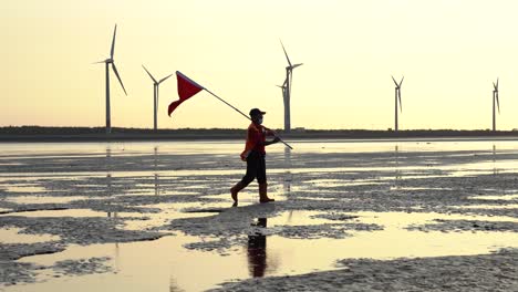 A-coast-guard-holding-a-red-flag,-walking-across-the-tidal-flats-with-wind-turbines-spinning-and-rotating-along-the-horizon-during-beautiful-sunset-at-golden-hours,-Gaomei-wetland,-Taichung,-Taiwan