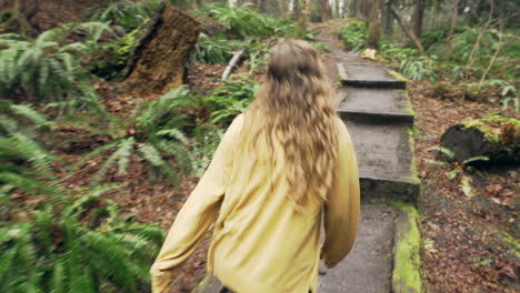 A-young-woman-wearing-a-bright-yellow-jacket-walks-over-a-small-bridge-in-a-green-mossy-forest,-tracking-shot-from-behind