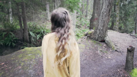 A-young-woman-wearing-a-bright-yellow-jacket-walks-over-a-small-bridge-in-a-forest,-tracking-shot-from-behind