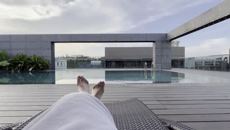 Someone-resting-on-the-poolside-outdoor-lounger-overlooking-at-calm-rooftop-pool-on-a-beautiful-sunny-day,-point-of-view-holiday-vacation-shot