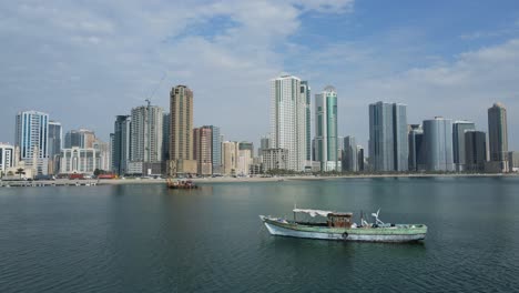 Drone-view-of-the-traditional-Arabian-fishing-boats-moored-on-the-surface-of-Gulf-Sea,-Mordren-skyscrapers-in-the-background