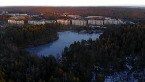 Luftaufnahme-Von-Menschen,-Die-Bei-Sonnenuntergang-Auf-Einem-Zugefrorenen-See-In-Einer-Von-Wald-Umgebenen-Stadt-Eislaufen