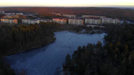 Aerial-Shot-Of-People-Ice-Skating-On-A-Frozen-Lake,-Beautiful-Village-In-A-Winter-Scene-At-Sunrise