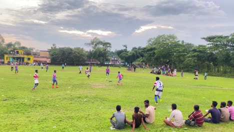 Young-men-in-Bangladesh-playing-football-on-grass,-Crowd-audience-watching-sitting-beside-the-field