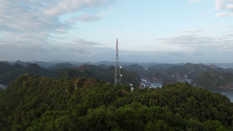 Vista-Aérea-Hacia-La-Torre-De-Comunicación-En-La-Cima-De-Una-Colina-Boscosa,-Bahía-De-Halong-En-El-Fondo,-Vietnam