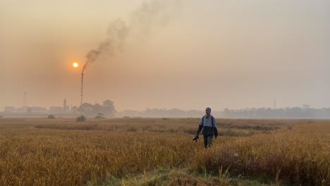 Photographer-walking-by-camera-in-vast-paddy-field-with-Gas-Burning-Plant-behind