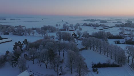 Aerial-establishing-shot-of-a-rural-landscape,-countryside-houses,-agricultural-fields-and-trees-covered-with-snow,-cold-freezing-weather,-sunset-golden-hour-light,-wide-drone-shot-moving-forward