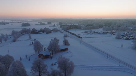 Aerial-establishing-shot-of-a-rural-landscape,-countryside,-agricultural-fields-and-trees-covered-with-snow,-cold-freezing-weather,-golden-hour-light-glow,-wide-drone-shot-moving-backward