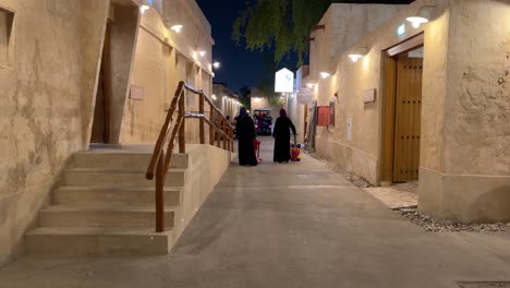 a-family-women-children-kids-are-walking-in-the-old-city-down-town-of-Doha-Qatar-a-walk-way-with-cobblestone-mudbrick-building-house-adobe-design-wooden-door-and-warm-light-offer-traditional-culture