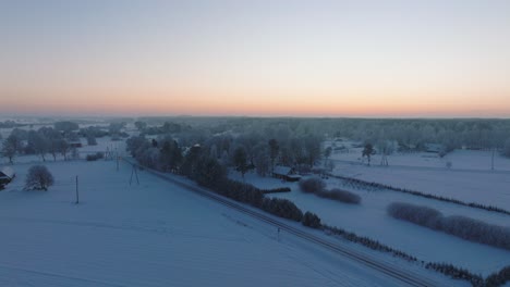 Aerial-establishing-shot-of-a-rural-landscape,-countryside-houses,-agricultural-fields-and-trees-covered-with-snow,-cold-freezing-weather,-sunset-golden-hour-light-glow,-wide-drone-shot-moving-forward