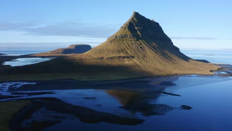 Rising-aerial-view-across-Kirkjufell-mountain-reflecting-in-calm-blue-sunset-waters-in-Snæfellsnes-peninsula,-Iceland